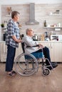Wife pushing invalid senior dreamy man in wheelchair in kitchen