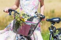 Wild flowers bouquet in basket of bicycle with woman in flowery dress in background, bike with flowers in field in summertime sunn