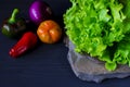 A wild multi-colored stone with a place for text lies on a black table next to fresh blue onions, yellow peppers, chili and