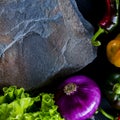 A wild multi-colored stone with a place for text lies on a black table next to fresh blue onions, yellow peppers, chili and