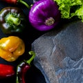 A wild multi-colored stone with a place for text lies on a black table next to fresh blue onions, yellow peppers, chili and