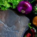 A wild multi-colored stone with a place for text lies on a black table next to fresh blue onions, yellow peppers, chili and