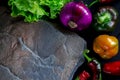 A wild multi-colored stone with a place for text lies on a black table next to fresh blue onions, yellow peppers, chili and