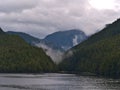 Landscape with cloud rising from tree-covered mountains at the shore of Tolmnie Channel, part of the Inside Passage, Canada.