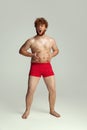 Cute red-headed man in red swimming shorts posing isolated on gray studio background. Concept of sport, humor and body