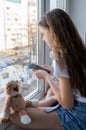 On the windowsill sits a cute teenage girl with a soft toy and a phone. The sun is shining outside the window.