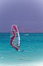 Windsurfer man on board with red colored sail on ocean on sunny day on blue sky background. Recreational water sports during idyll