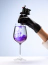 Wine glass and purple ink. Tattooer master& x27;s hand in black glove holding machine for making tattoo art on body