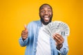 Winner! Young rich african american man in casual t-shirt holding money dollar bills with surprise isolated over yellow wall.