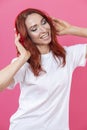 Winsome cheerful stylish young woman wearing white shirt standing isolated over pink background while listening to music with Royalty Free Stock Photo