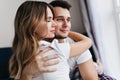 Winsome girl with long hair embracing her boyfriend with eyes closed. Indoor portrait of cute married couple spending