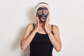 Winsome girl treating and caring for her skin. Woman with hairband on head and facial mask looking at camera over white background