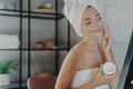 Winsome pleased young European woman puts cream on face, looks at her reflection in mirror, wrapped in bath towel, has anti aging Royalty Free Stock Photo