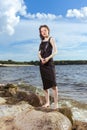 Winsome Relaxing Caucasian Brunette Girl Posing in Black Dress On Stone Line At Sea During Sunny Day Outdoors