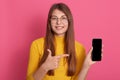 Winsome young beautiful woman shows to camera blank screen of smart phone, pointing at cellphone with index finger isolated over Royalty Free Stock Photo