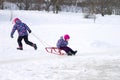 Elder girl running and pulling her young sister on a sled on the ice in snowy winter park Royalty Free Stock Photo