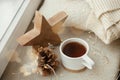 Winter and autumn hygge. Stylish cup of tea with cozy knitted sweater, pine cone, wooden star and golden lights on windowsill in