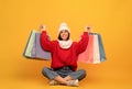 Winter big sale. Excited young woman in white hat and knitted sweater with shopping bags, sitting over yellow background