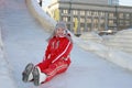 In winter, a cheerful girl in a red jumpsuit a gray cap with a earflap rolls off an ice slide, rides sitting with stretched legs Royalty Free Stock Photo