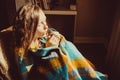 Winter cold concept. Young freezing woman in comfortable chair warming frozen hands wrapped in warm fluffy woollen plaid blanket.