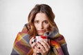 Winter cold sickness concept. Freezing beautiful woman wrapped in warm checkered plaid blanket, drinks hot beverage, tries to warm Royalty Free Stock Photo