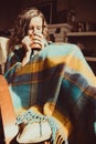 Winter cold sickness concept. Young freezing woman sitting in comfortable modern chair with mug of tea wrapped in warm plaid blank
