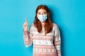 Winter, covid-19 and quarantine concept. Excited redhead woman in sweater and face mask, pointing finger at upper left