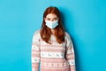 Winter, covid-19 and quarantine concept. Young redhead girl in sweater and face mask staring at camera, standing over