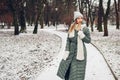 Winter fashion. Portrait of young woman wearing long green coat with scarf, hat, mittens in snowy park. Royalty Free Stock Photo