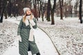 Winter fashion. Portrait of young woman wearing long green coat with scarf, hat, mittens in snowy park Royalty Free Stock Photo