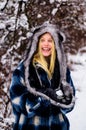 Winter fashion. Smiling girl in warm plaid coat, fur hat and mittens in snowy winter park. Blonde woman in fashionable Royalty Free Stock Photo