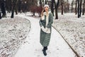 Winter fashion. Young woman wearing long green coat with scarf, hat, mittens and boots in snowy park. Royalty Free Stock Photo