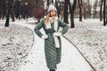 Winter female fashion. Young woman wearing long green coat with scarf, hat, mittens in snowy park. Royalty Free Stock Photo