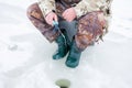 Winter fishing on ice. Fisherman in camouflage uniforms sitting on a box on ice, holding a fishing ice rod for winter fishing, low