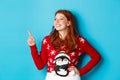 Winter holidays and Christmas Eve concept. Happy and cute redhead girl in xmas sweater, looking and pointing at upper