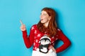 Winter holidays and Christmas Eve concept. Happy and cute redhead girl in xmas sweater, looking and pointing at upper