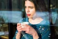 Winter holidays, christmas, hot drinks concept. Happy beautiful young woman drinking coffee behind a window Royalty Free Stock Photo