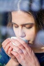 Winter holidays, christmas, hot drinks and people concept. Happy beautiful young woman drinking coffee behind a window Royalty Free Stock Photo