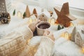 Winter hygge. Hands holding stylish cup of tea with modern cute christmas houses, pine cone, wooden star and tree, golden lights Royalty Free Stock Photo