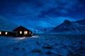 Winter image of a wooden house with light from the window. Landscape with moonlight on the snow with mountains in the background.