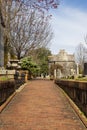 A winter landscape along a red brick footpath with bare winter trees, lush green plants with flowers headstones and graves