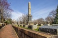 A winter landscape along a red brick footpath with bare winter trees, lush green plants with flowers headstones and graves