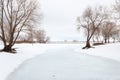 A winter landscape of a frozen river going to the pespective far away. Leafless trees are on the sides of the frame and in the