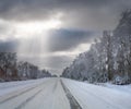 Winter landscape with road, trees at side of the road, and sunrays from cloudy sky Royalty Free Stock Photo