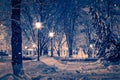 Winter night park with lanterns, pavement, fountain and trees covered with snow in snowfall Royalty Free Stock Photo