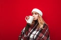 Winter portrait of cute blonde in plaid and hat santa stands on red background and drinks tea from big cup and looks aside at copy Royalty Free Stock Photo