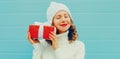 Winter portrait happy smiling woman holding red gift box in her hands wearing a white knitted hat, sweater on blue background Royalty Free Stock Photo