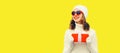 Winter portrait of happy smiling woman holding red gift box in her hands wearing white knitted hat, sweater on yellow background, Royalty Free Stock Photo