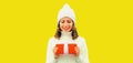 Winter portrait of happy smiling woman holding red gift box in her hands wearing white knitted hat, sweater on yellow background, Royalty Free Stock Photo