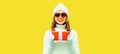 Winter portrait of happy smiling woman holding red gift box in her hands wearing white knitted hat, sweater on yellow background, Royalty Free Stock Photo